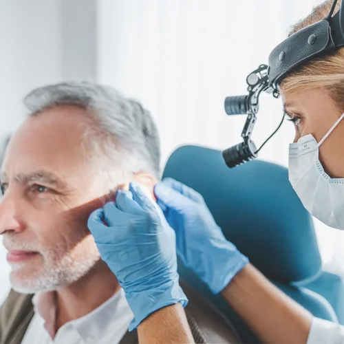 Audiologist wearing a magnifying headlamp examining an older man’s ear